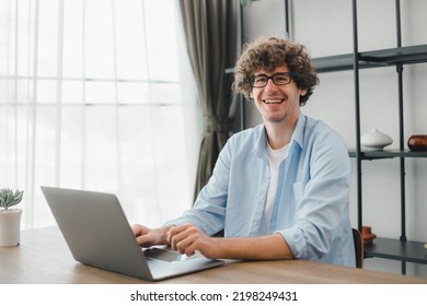 Happy Young Caucasian Man Wearing Glasses And Smiling Working Typing On His Laptop To Get All His Business Done Early In The Morning With His Cup Of Coffee. Young White Man Studying Online At Home.