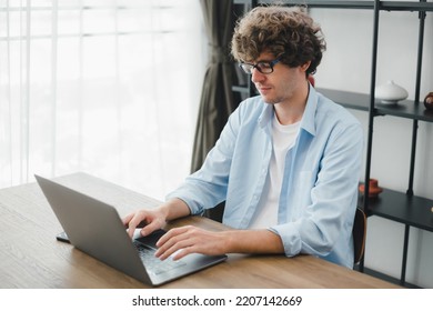 Happy Young Caucasian Man Wearing Glasses And Smiling Working Typing On His Laptop To Get All His Business Done Early In The Morning With His Cup Of Coffee. Young White Man Studying Online At Home.