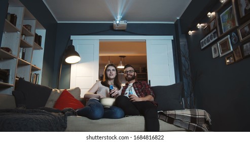 Happy Young Caucasian Man And Woman Sit Together Watching Fun Movie At Home With Snacks Using Projector Slow Motion.