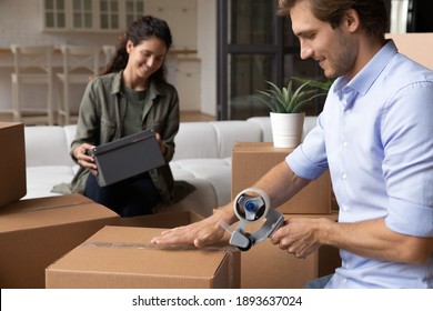 Happy young Caucasian man and woman seal wrap packages with tape dispenser relocate to new house. Smiling millennial couple renters pack boxes with adhesive scotch, moving to own home together.