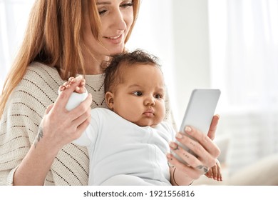 Happy Young Caucasian Mother Holding Cute African American Baby Girl Looking At Smartphone At Home. Diverse Mum And Ethnic Little Infant Child Daughter Using Apps On Mobile Cell Phone Device.