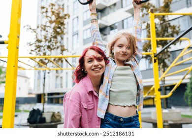 Happy young Caucasian mother supporting her teen daughter while playing with the monkey bars in jungle gym on playground with modern building on background. - Powered by Shutterstock