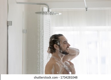 Happy young caucasian naked man stand under shower in home bathroom washing head body in morning, millennial male enjoy taking bath cleaning cleansing with foamy shampoo or gel, hygiene concept