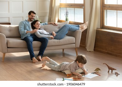 Happy Young Caucasian Parents Relax On Cozy Couch In Living Room At Home. Small 7s Son Lying On Warm Floor Drawing Painting In Album. Overjoyed Family With Little Child Rest On Weekend In Own House.