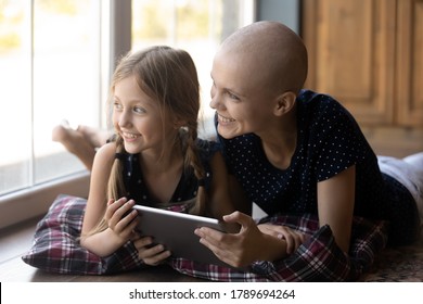 Happy Young Caucasian Sick Cancer Patient Bald Hairless Mom And Little Daughter Rest On Floor Use Tablet Look In Window Distance Dreaming, Smiling Ill Mother And Small Girl Child Relax With Pad Gadget