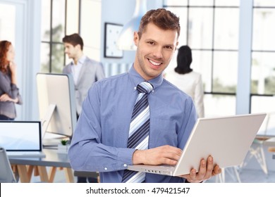 Happy Young Caucasian Stock Broker At Business Office With Laptop Computer In Hand. Standing, Smiling, Looking At Camera. Tie, No Jacket.