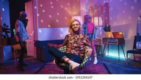 Happy young Caucasian stylish guy sitting in chair in living room in neon disco ball light, looking at camera and smiling while multi-ethnic friends dancing and moving behind having fun at party time