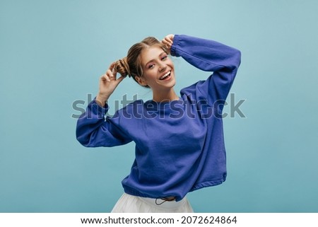 Happy young caucasian teenager looking at camera holding bundles of hair on her head indoors. Blonde with bright makeup is cheerfully posing smiling broadly, dressed in dark blue body shirt. 