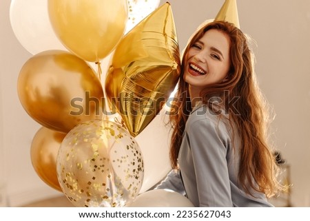 Happy young caucasian woman with balloons laughing looking at camera indoors. Long-haired redhead lady wears pajamas at bachelorette party. Holiday concept