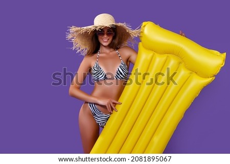 Happy young caucasian woman in bikini in straw hat in sunglasses holding yellow swimming mattress while isolated on purple background 