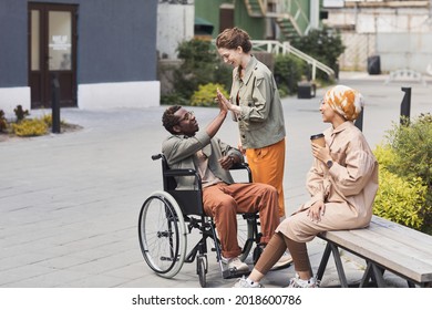 Happy Young Caucasian Woman Giving Five To Disabled Black Man In Wheelchair While Welcoming Him Outdoors