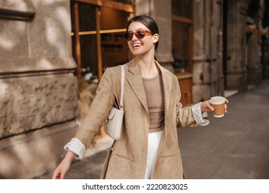 Happy young caucasian woman in good mood walks with coffee around city during day. Brunette wears sunglasses, jacket and bag. Real emotions concept