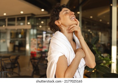 Happy young caucasian woman laughs with eyes closed and head tilted back standing indoors. Brunette is wearing white sweatshirt over her shoulders. Concept of enjoying moments
