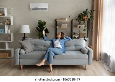 Happy Young Caucasian Woman Relax On Couch In Living Room Turn On Air Conditioner With Remote Controller. Smiling Female Rest On Sofa At Home Breathe Fresh Air From Ac Electronic Condition Device.