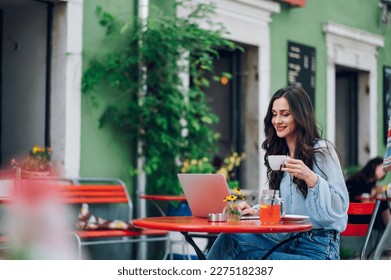 Happy young caucasian woman relaxing in outdoor cafe and drinking coffee while working on a laptop. Portrait of a businesswoman working outside during a sunny day. Copy space. - Powered by Shutterstock