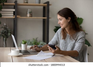 Happy young Caucasian woman sit at desk look at laptop screen use cellphone study online from home, smiling female work on computer or take web course, make notes summarize, distant education concept