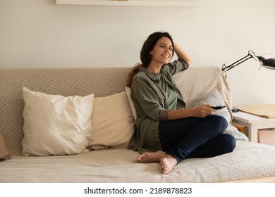 Happy Young Caucasian Woman Sit Relax On Comfortable Couch In Living Room Watching TV Programs. Smiling Millennial Female Rest On Sofa At Home Enjoy Prepaid Satellite Television On Leisure Weekend.