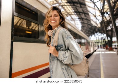 Happy young caucasian woman smiles with teeth looking at camera going on train trip. Blonde wears denim jacket, backpack. Spring break concept. - Powered by Shutterstock