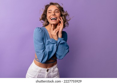 Happy young caucasian woman smiles broadly with teeth looking away on purple background. Blonde girl with wavy hair wears blouse and pants. Concept of human emotions - Powered by Shutterstock