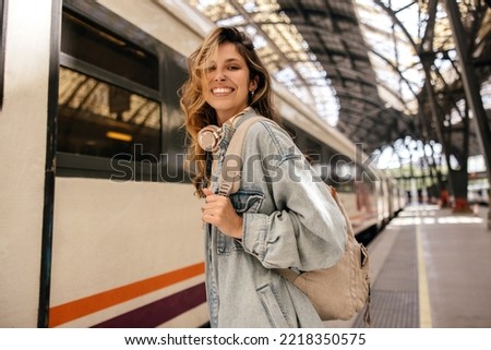 Happy young caucasian woman smiles with teeth looking at camera going on train trip. Blonde wears denim jacket, backpack. Spring break concept.
