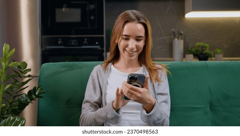 Happy young caucasian woman using mobile phone while sitting a green couch at home kitchen background. Smiling girl with blond hair relaxing on cozy sofa and browsing internet on smartphone
