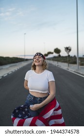 A Happy Young Caucasian Woman Wearing A Bandana And Holding A US Flag While Walking On The Road