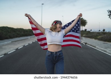 A Happy Young Caucasian Woman Wearing A Bandana And Holding A US Flag While Walking On The Road