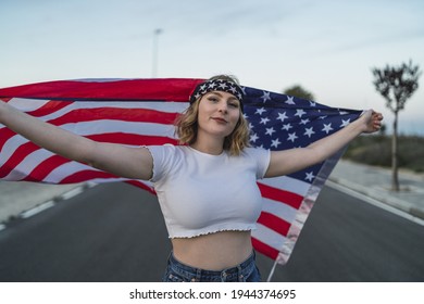 A Happy Young Caucasian Woman Wearing A Bandana And Holding A US Flag While Walking On The Road