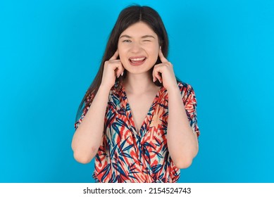 Happy Young Caucasian Woman Wearing Floral Dress Over Blue Background Ignores Loud Music And Plugs Ears With Fingers Asks To Turn Off Sound