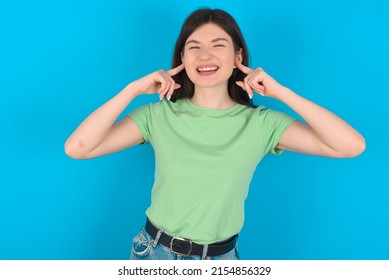 Happy Young Caucasian Woman Wearing Green T-shirt Over Blue Background Ignores Loud Music And Plugs Ears With Fingers Asks To Turn Off Sound