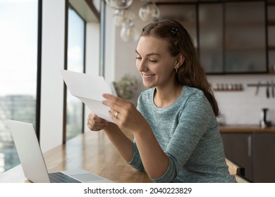 Happy young Caucasian woman work on laptop at home office read good news in paperwork correspondence. Smiling millennial female employee get pleasant message or promotion offer in letter. - Shutterstock ID 2040223829
