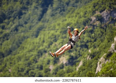 Happy young cheerful female tourist wearing casual clothes riding on zipline in forest.  Zipline trip selective focus against blurred background. Holiday, adventure, extreme sport concept.