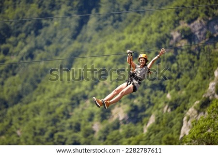 Happy young cheerful female tourist wearing casual clothes riding on zipline in forest.  Zipline trip selective focus against blurred background. Holiday, adventure, extreme sport concept.