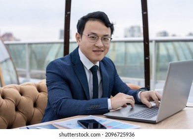 Happy Young Chinese Businessman In Formalwear And Eyeglasses Working In Front Of Laptop While Sitting By Table In Cafe