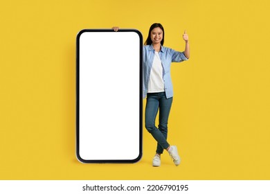 Happy young chinese female student in casual show thumb up, stand near huge smartphone with blank screen, isolated on yellow background, studio. App and technology for study and work, ad and offer - Shutterstock ID 2206790195