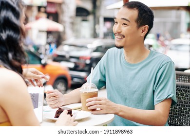 Happy Young Chinese Man Drinking Cold Coffee, Talking To Girlfriend And Taking Notes In Textbook