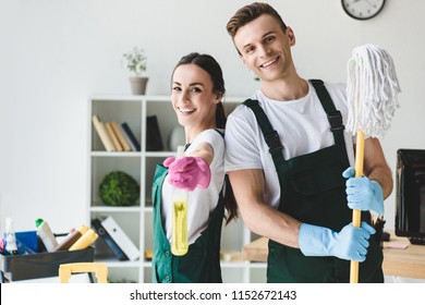 happy young cleaners with mop and spray bottle smiling at camera while cleaning office