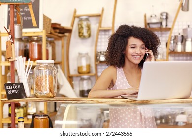Happy young coffee shop owner talking on he mobile phone while typing on her laptop on the counter