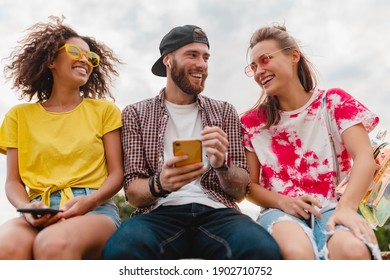 Happy Young Company Of Smiling Friends Sitting Park Using Smartphones, Man And Women Having Fun Together, Colorful Summer Hipster Fashion Style, Communication Wireless Connecting Devices