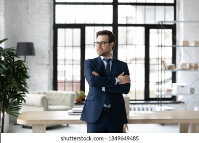 Happy young confident businessman in formal suit and glasses standing with folded arms in modern workplace, looking away, thinking of company development challenges or career opportunities indoors.