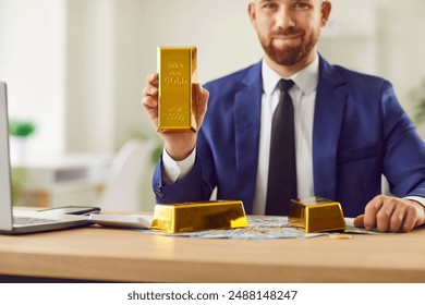 Happy young confident businessman in suit sitting at desk with money bills and laptop and looking cheerful at camera. Male person holding gold bar in hands. Investment and success concept - Powered by Shutterstock