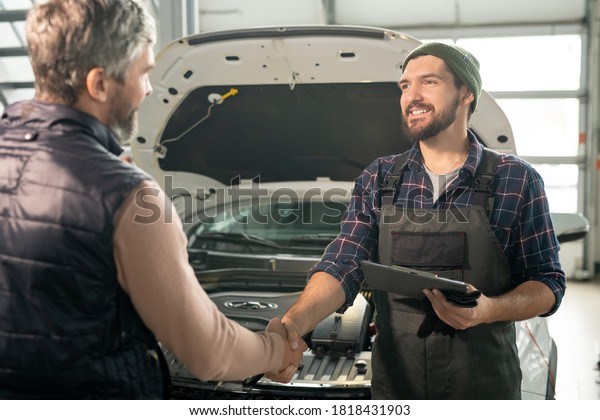 Happy young contemporary technician of car\
service shaking hand of male client against front part of broken\
car with open engine\
compartment