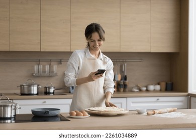 Happy young cook girl wearing apron, using mobile phone in home kitchen while baking pie, pizza, pastry, taking photo of raw bakery food for blog, chatting, making call - Powered by Shutterstock