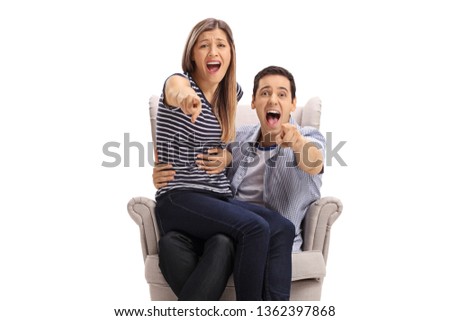 Happy young couple in an armchair pointing at the camera and laughing isolated on white background