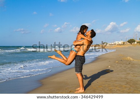 Happy young couple in bikini and shorts enjoying summer dusk at the beach, having fun walking barefoot, kissing and teasing one another.
