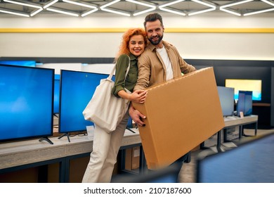 Happy Young Couple Bought New TV, Holding Box With Purchase In Store Of Household Appliances, Electronics And Gadgets. Buying New TV, Sale Day.