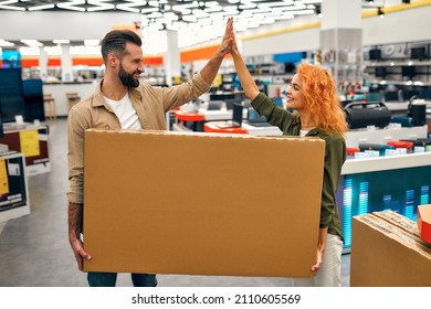 Happy Young Couple Bought A New TV, Holding A Box With A Purchase In A Home Appliances, Electronics And Gadgets Store And Giving A High Five Gesture. Buying A New TV, Sale Day.