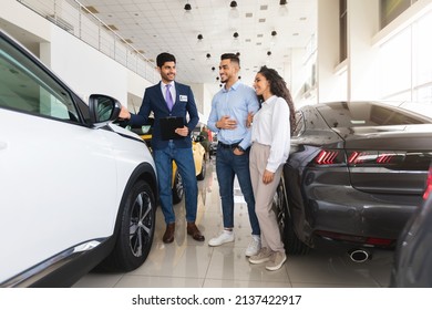 Happy Young Couple Buying New Auto At Car Showroom, Handsome Middle Eastern Man Sales Manager Assisting Wealthy Family, Standing By Nice White Automobile, Having Convesation, Full Length