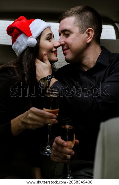 Happy young couple in a car. Man and woman\
celebrating Christmas with champagne in car. Woman wearing Santa\'s\
hat. Sensual lovers in black\
clothes.