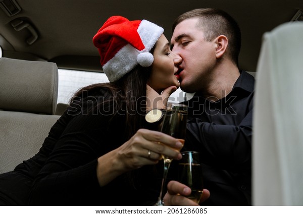Happy young couple in a car. Man and woman\
celebrating Christmas with champagne in car. Woman wearing Santa\'s\
hat. Sensual lovers in black\
clothes.
